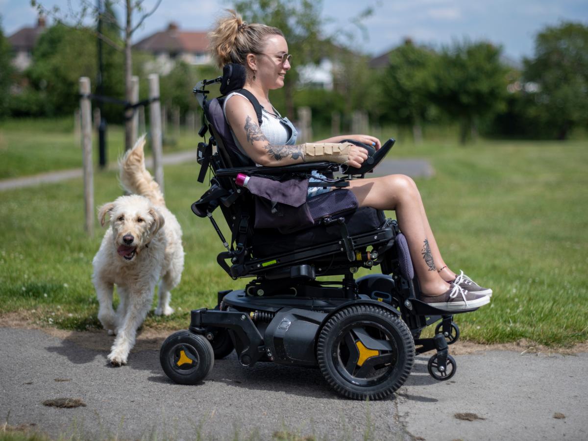 Young woman operating powered wheelchair in park with dog running behind her